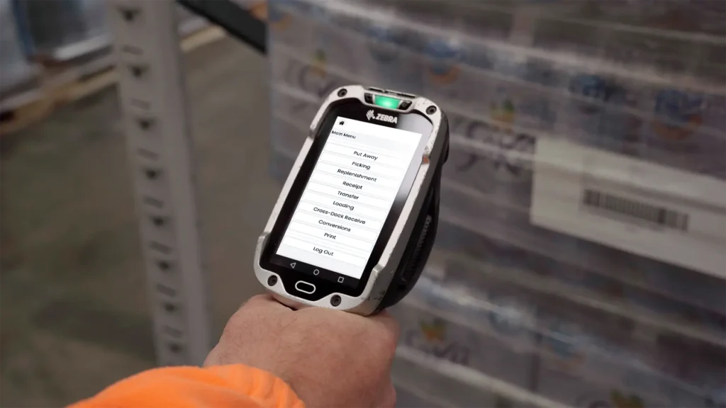 Hand holding a zebra scanner showing the clarus wms main menu with picking and put away options