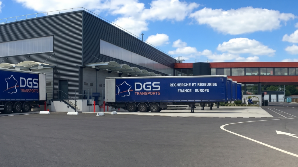 Dgs transports logistics facility | clarus wms Several blue dgs transports semi-trailers parked at the loading docks of a large, modern grey warehouse under a bright blue sky with white clouds.