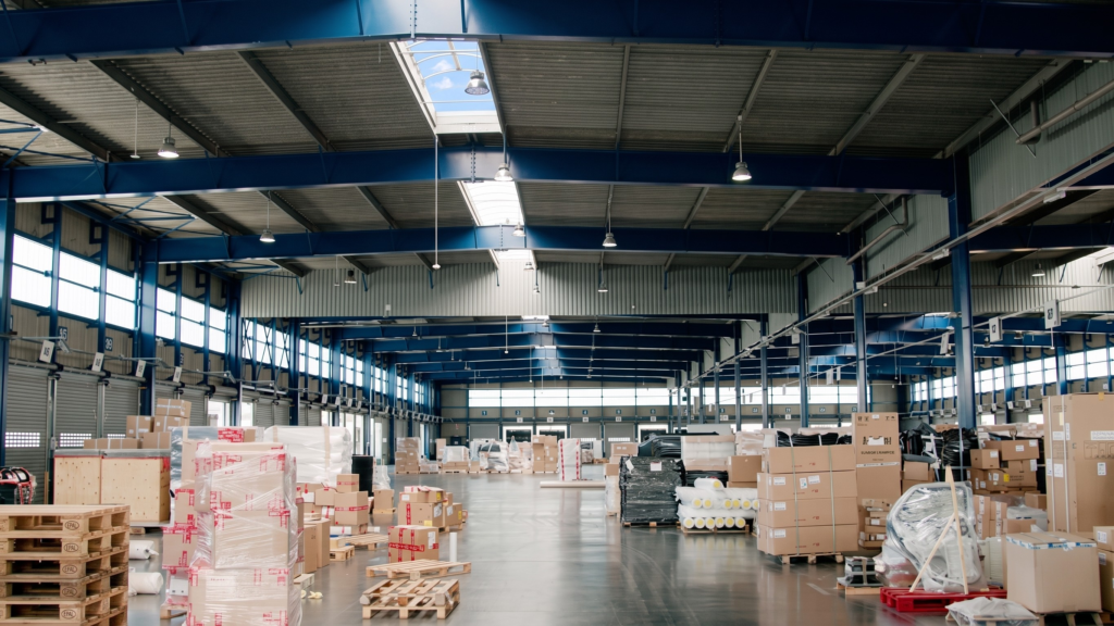 Spacious warehouse interior with cargo | clarus wms The interior of a large, brightly lit warehouse with a blue steel frame, showing various pallets of cardboard boxes and shrink-wrapped goods resting on a polished concrete floor.